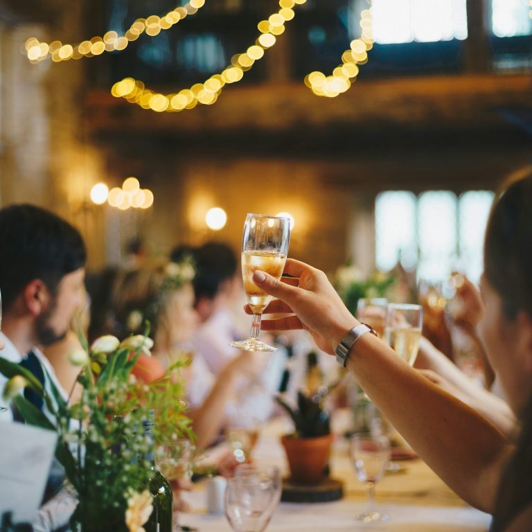 Gläser werden bei einer Hochzeit erhoben, im Hintergrund festliche Dekoration und Gäste.